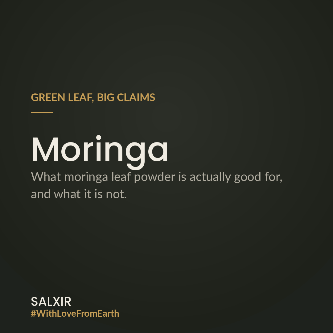 A spoonful of bright green moringa leaf powder beside dried moringa leaves on a pale stone surface